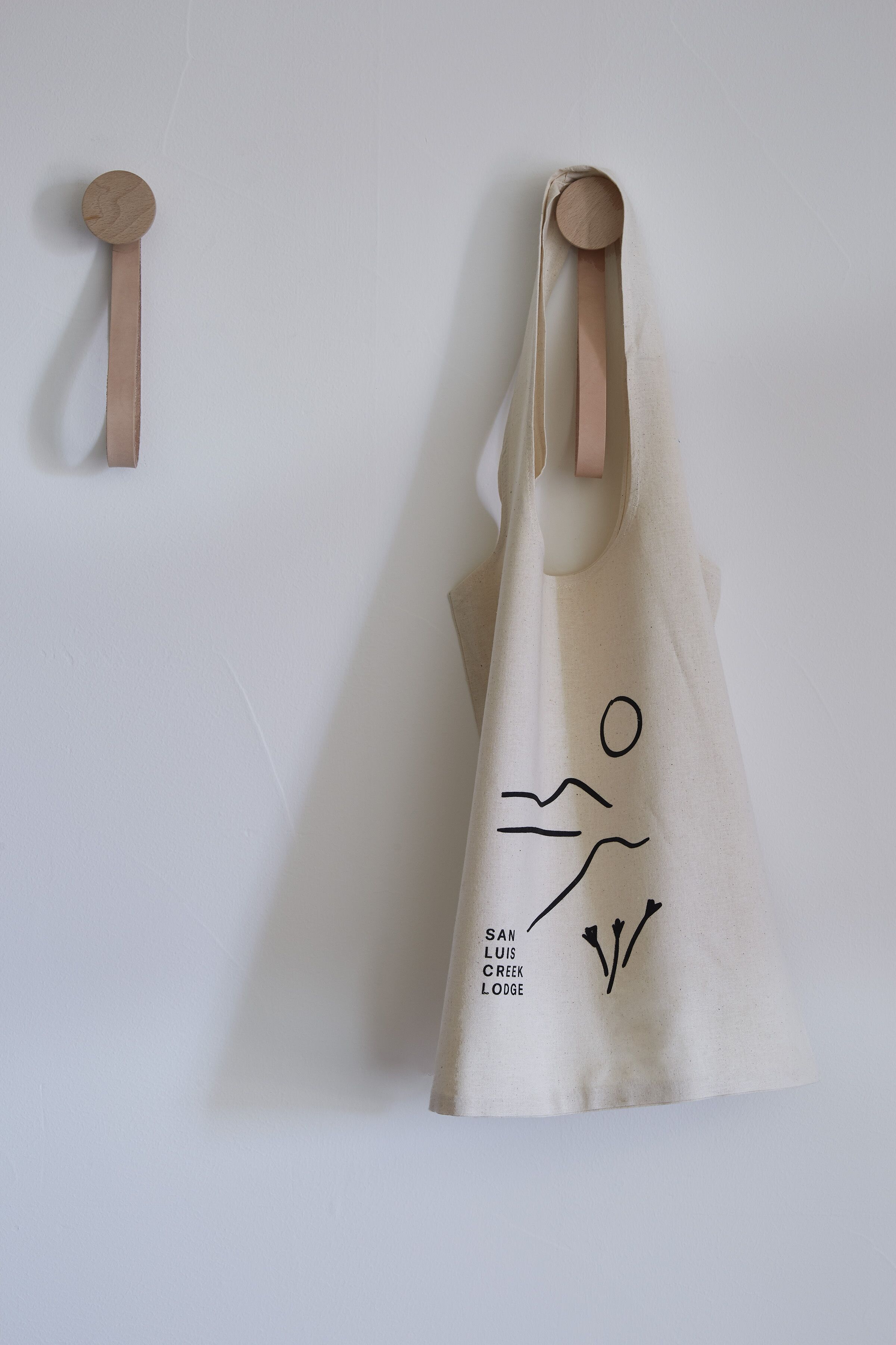 Un sac fourre-tout beige orné d’un simple dessin linéaire et d’un texte, accroché à un mur blanc.