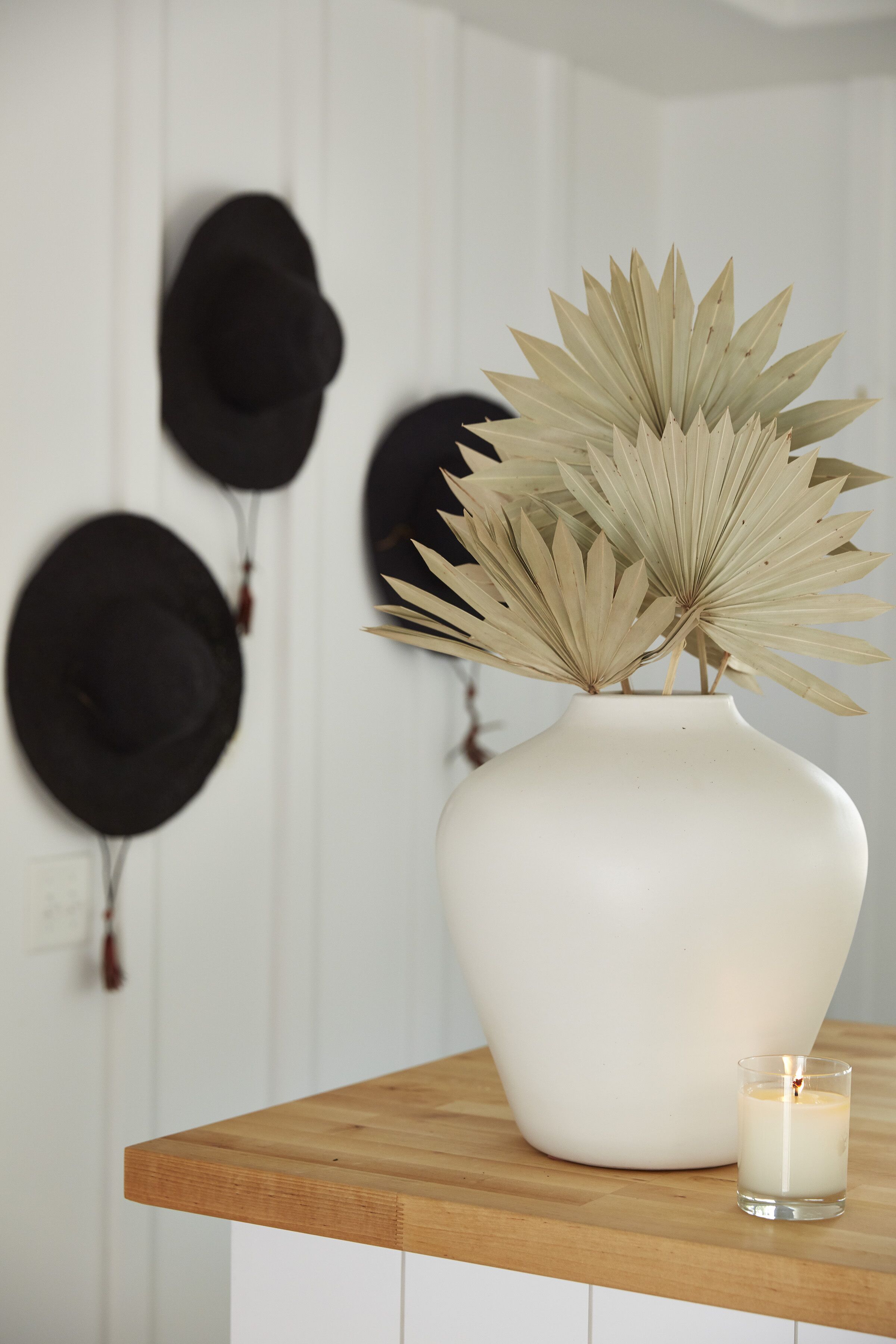 Un jarrón blanco con hojas secas de planta sobre una mesa de madera, rodeado de sombreros negros en una pared.