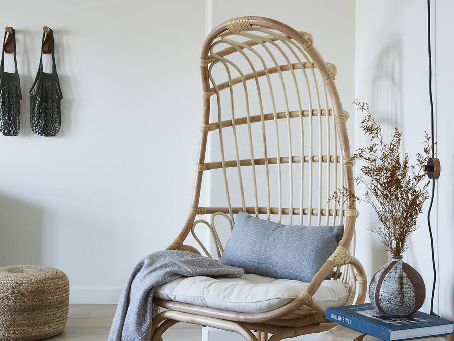 A wicker chair with a cushion, a small wooden side table with a book, and a black floor lamp.