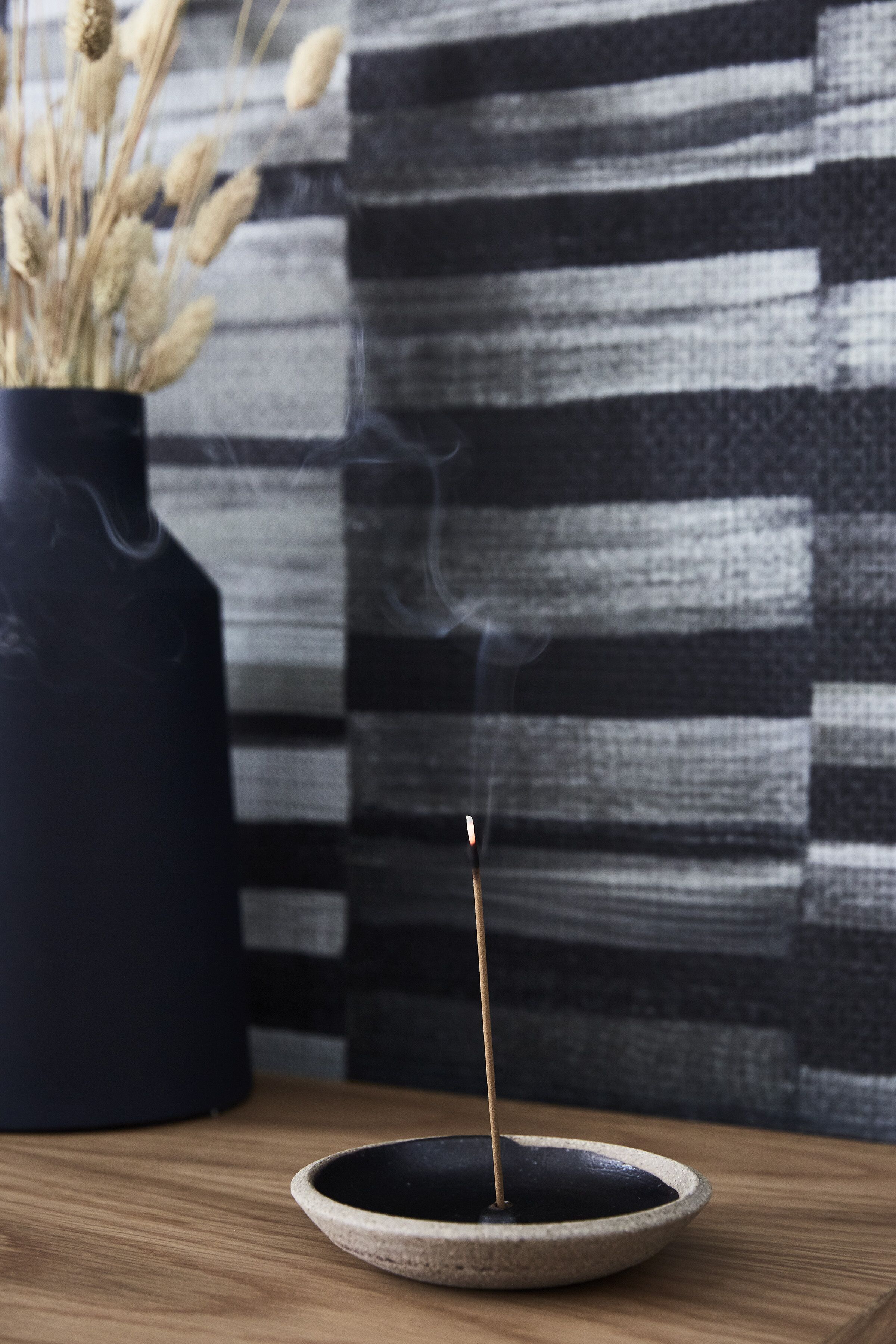 A candle in a ceramic bowl on a wooden surface, with a striped fabric background and a dark vase with dried plants to the left.