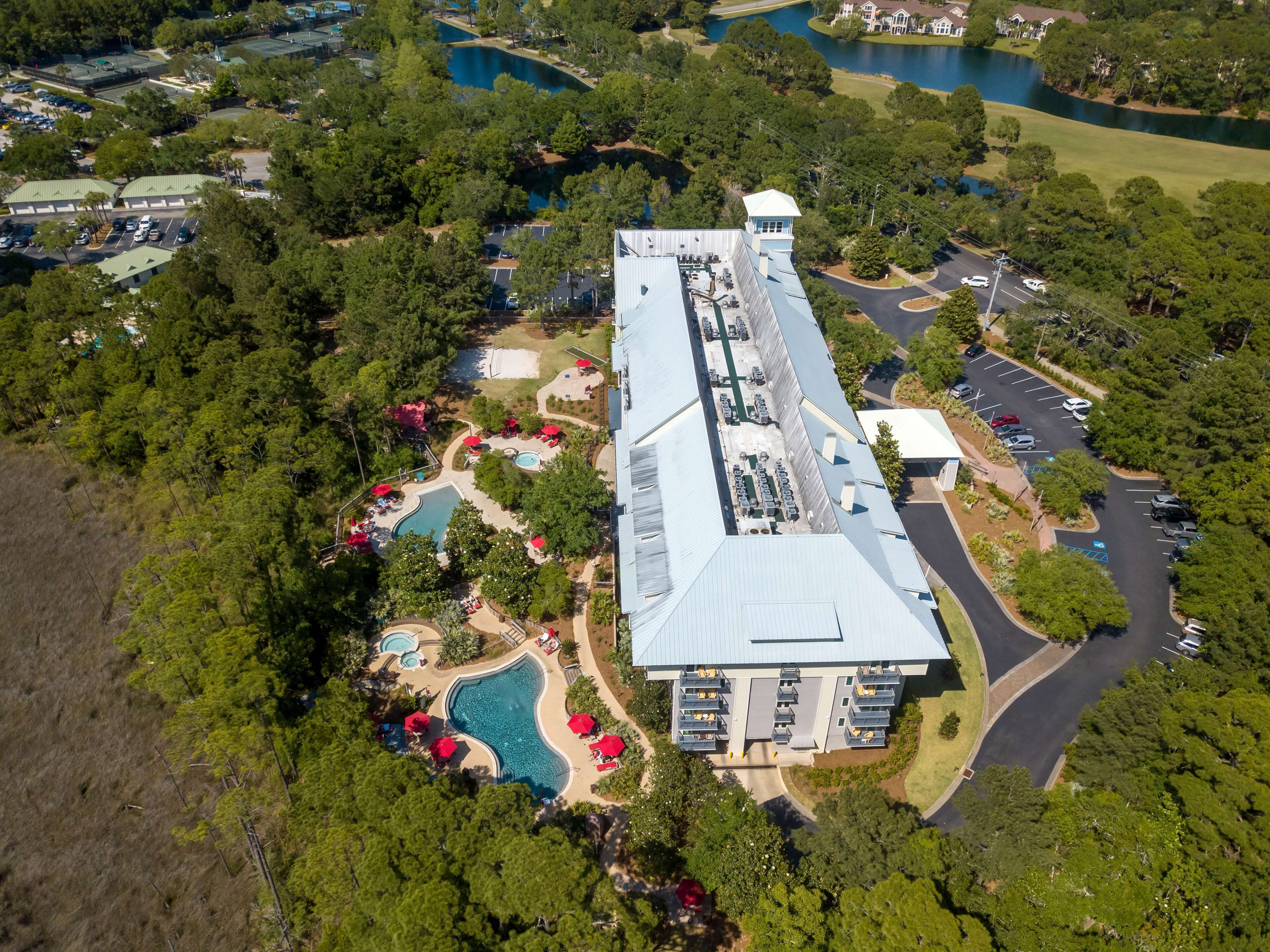 Ovenudblik over et hotel med swimmingpool, tennisbaner og en stor parkeringsplads.