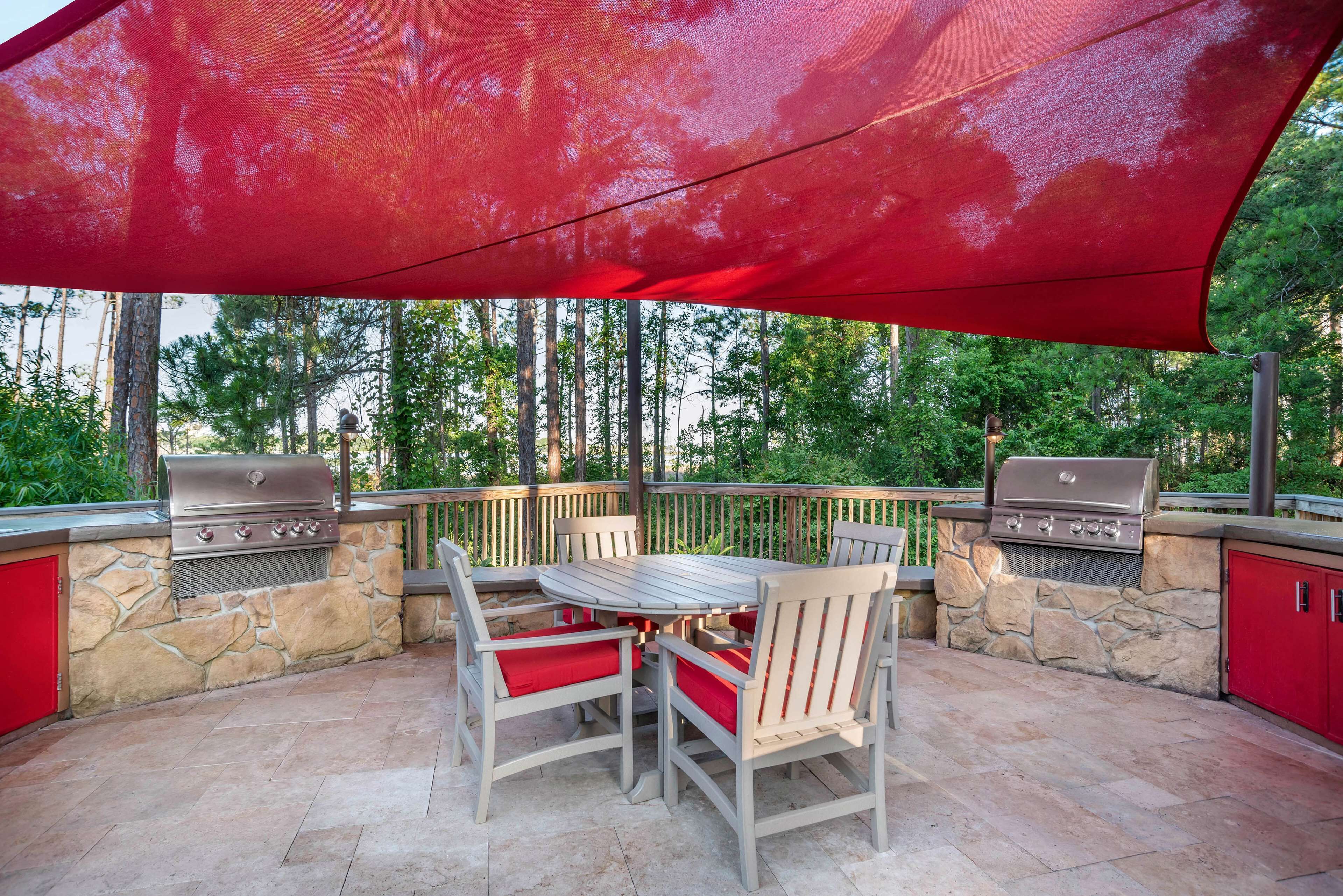 An outdoor patio with a stone wall, two barbecue grills, a round table with chairs, and a red canopy.