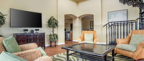 A spacious living room with a flat-screen TV, wicker furniture, a coffee table, and a staircase with ornate railings.