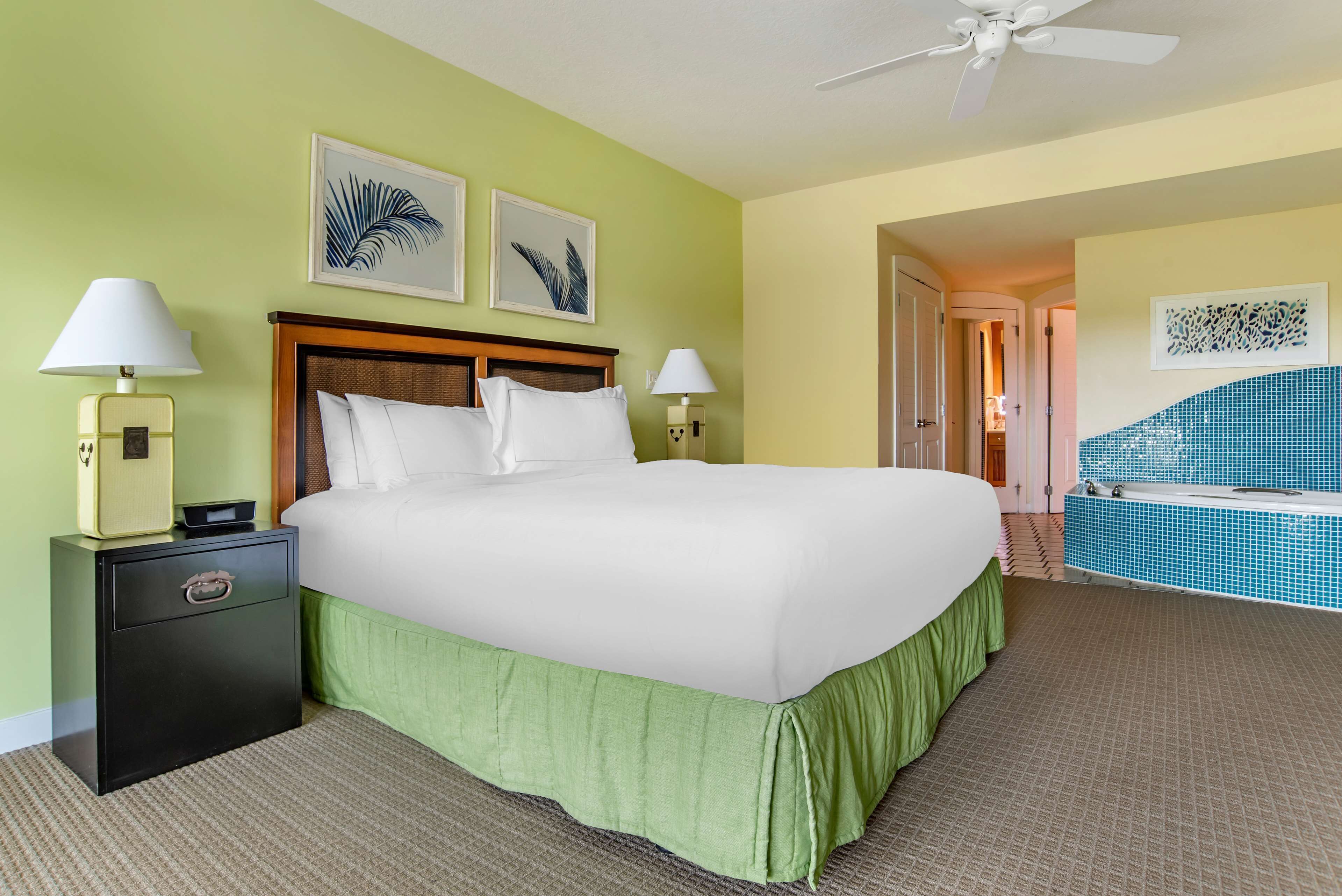 A hotel room with a large bed, two bedside tables with lamps, a ceiling fan, and a view of a pool area.