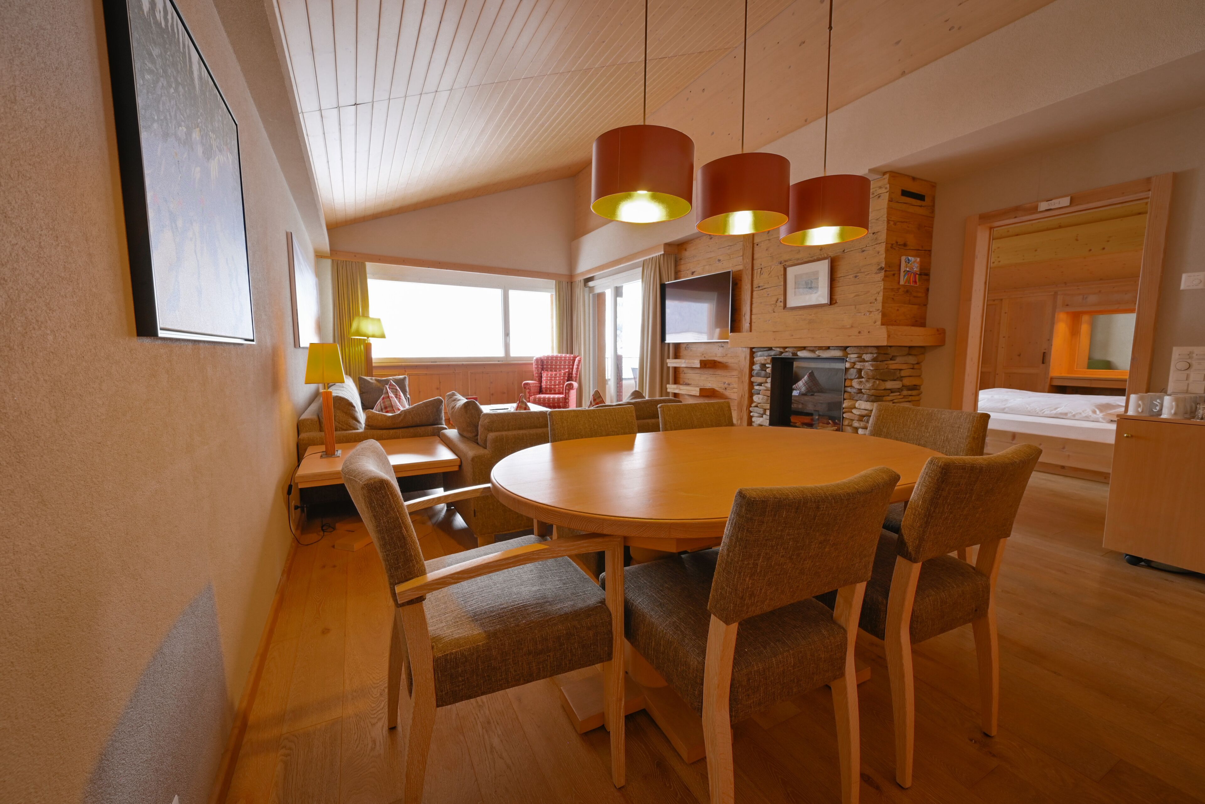 A spacious dining area with a round wooden table and six upholstered chairs. There are two pendant lights above the table, and a television is mounted on the wall.