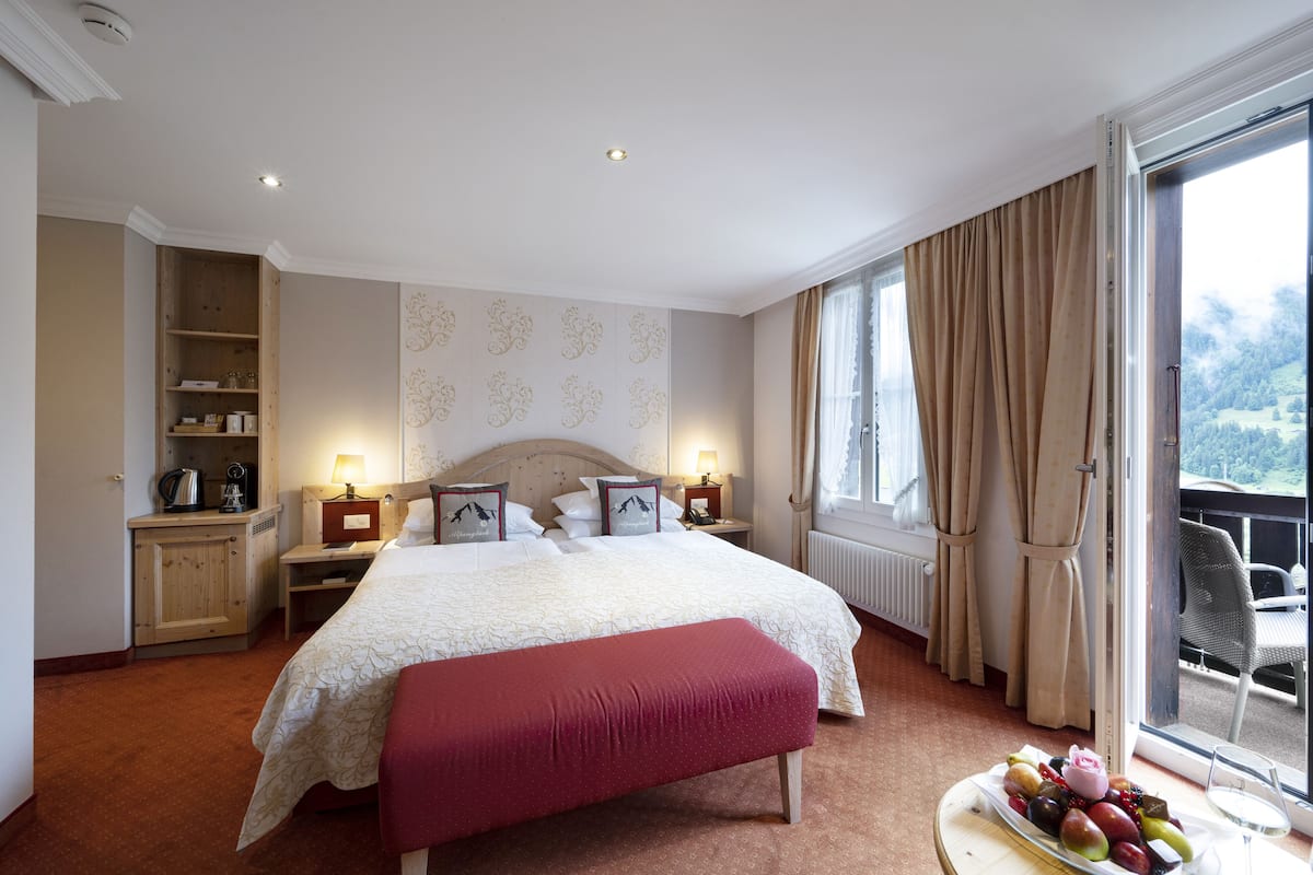 Habitación de hotel con una cama grande, un armario de madera, una mesita de noche con lámpara, una ventana con cortinas y un balcón con silla.