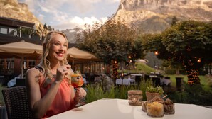 Una persona sentada en una mesa al aire libre con montañas de fondo, sosteniendo una bebida.