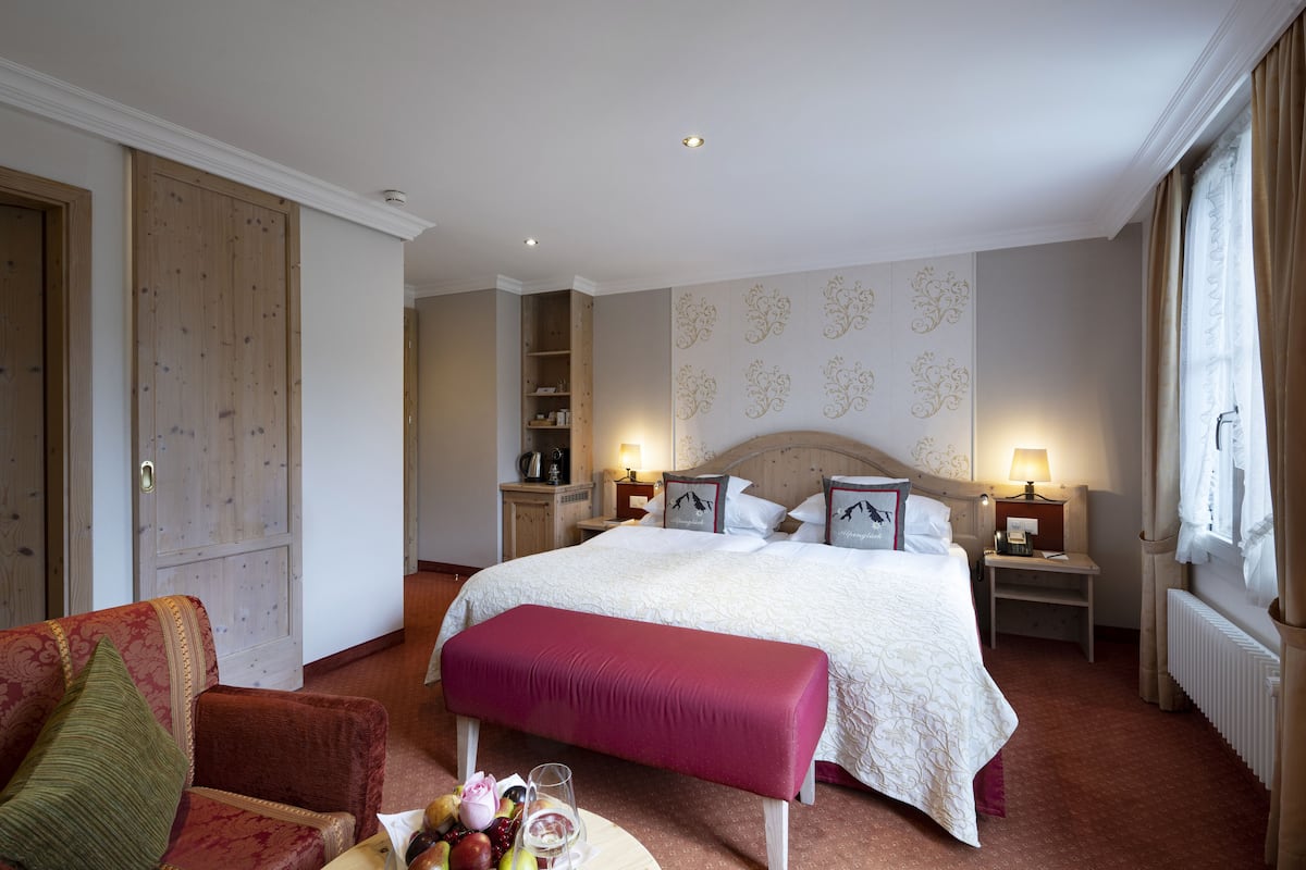 Una habitación de hotel con una cama grande, un rincón de descanso con una silla, una mesita con fruta y un vaso, y un armario.