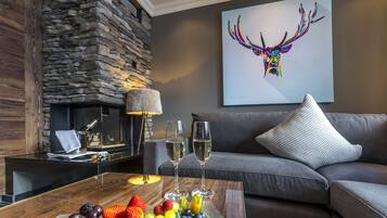 A cozy living room with a stone fireplace, a grey sofa, a coffee table with fruit and two glasses of wine, and a colorful deer painting on the wall.