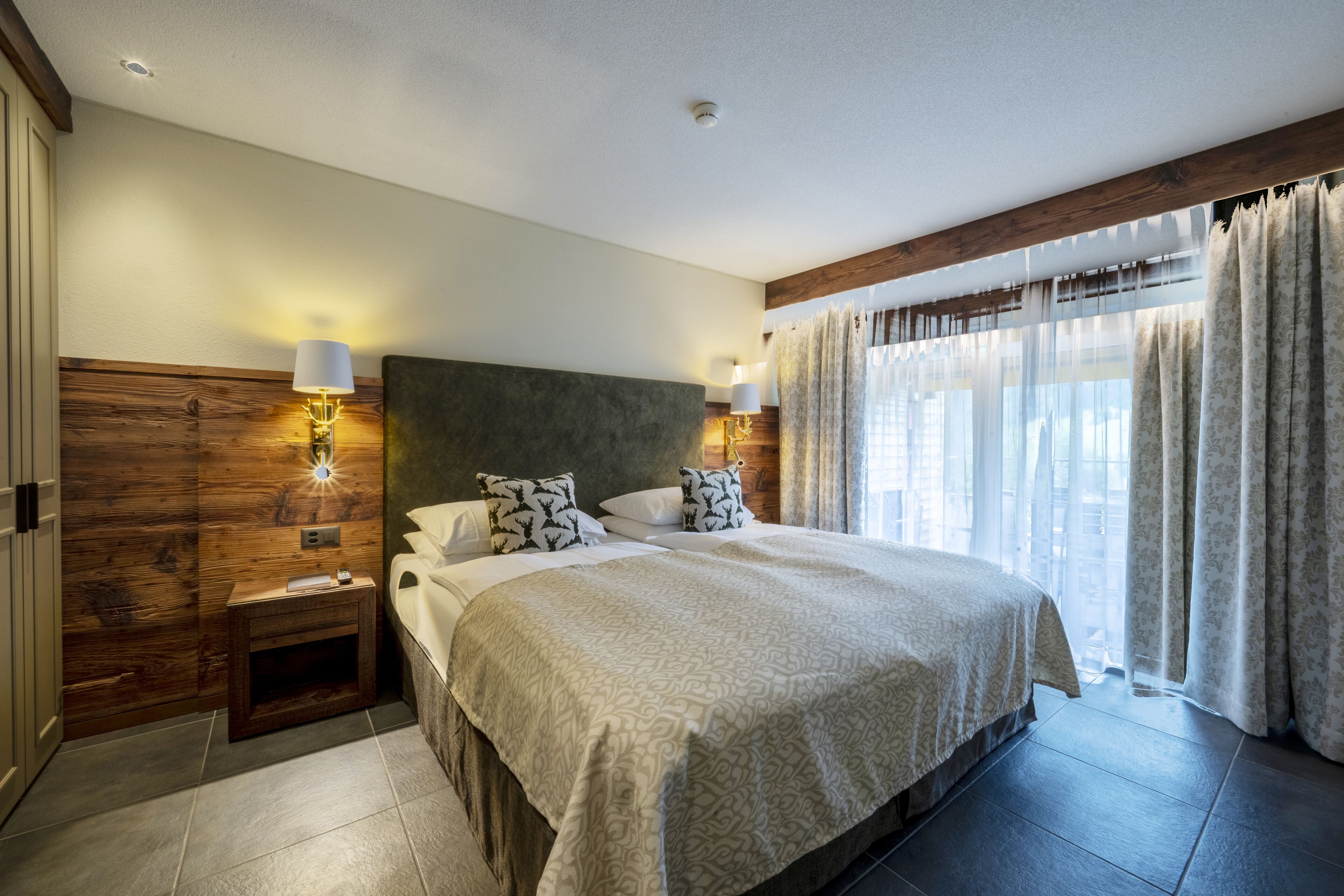 Une chambre à coucher avec un grand lit, une tête de lit en bois, des lampes de chevet et une vue sur l’extérieur à travers des rideaux légers.