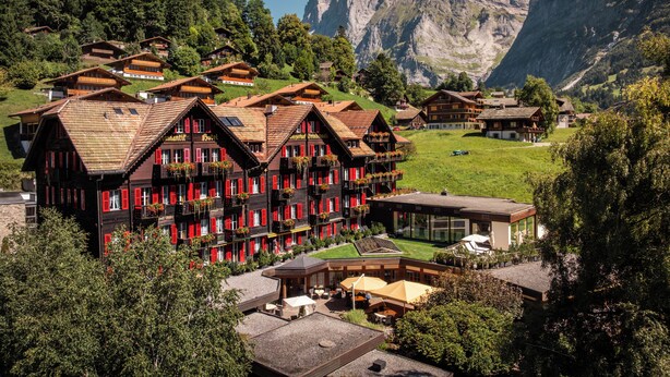 Un hotel de estilo chalete tradicional, con varios balcones, un telón de fondo montañoso y un jardín cuidado.