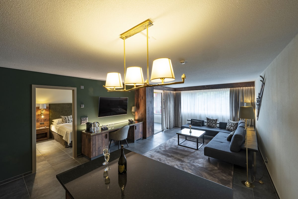 Una habitación de hotel moderna con sala de estar, una mesa de comedor con una botella de vino, un sofá y un televisor.