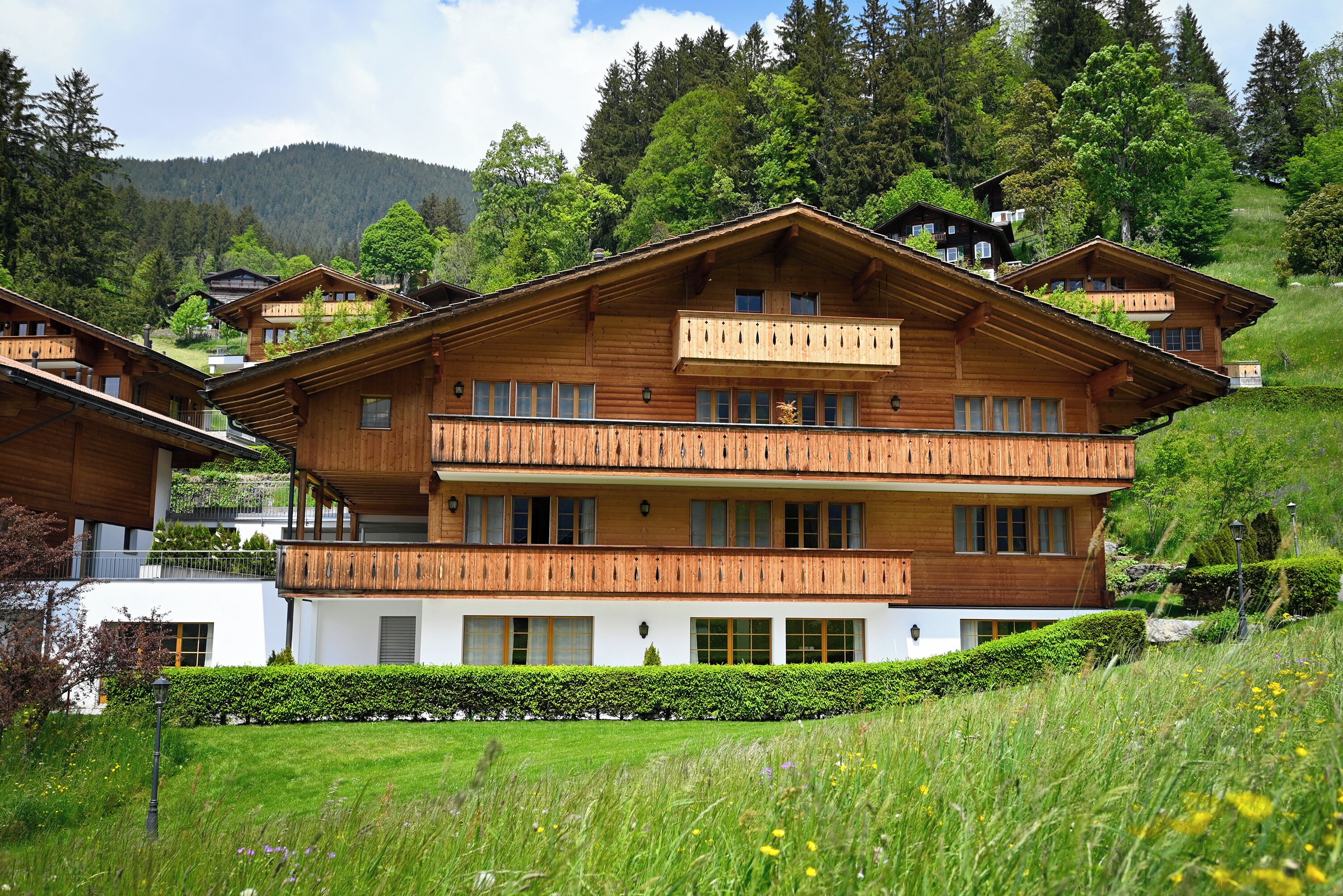 A multi-story wooden chalet with balconies, surrounded by lush greenery and mountains in the background.