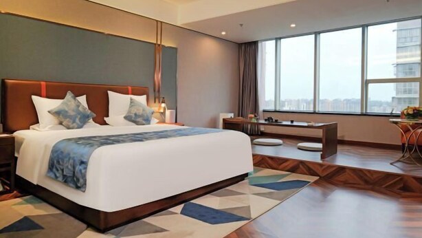 A modern hotel room with a large bed, a desk, and a view of the city through large windows. - Mingxing Conifer Hotel (Suining)