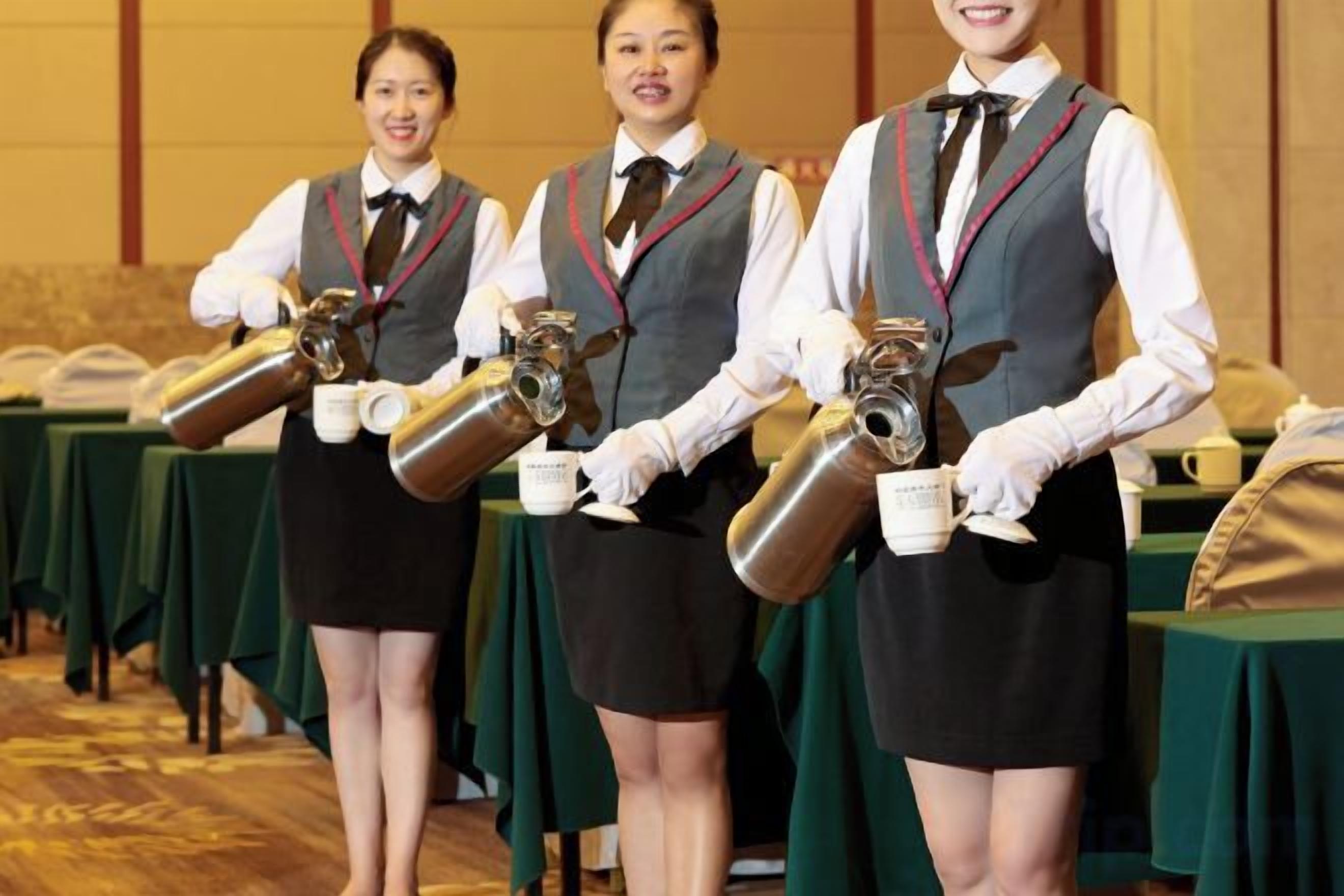 Tres camareras sostienen grandes recipientes metálicos, probablemente para bebidas, en una sala con mesas cubiertas con manteles verdes.