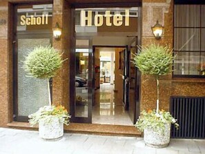 A hotel entrance with a sign reading "Hotel" and "Scholl". There are two potted plants on either side of the entrance.