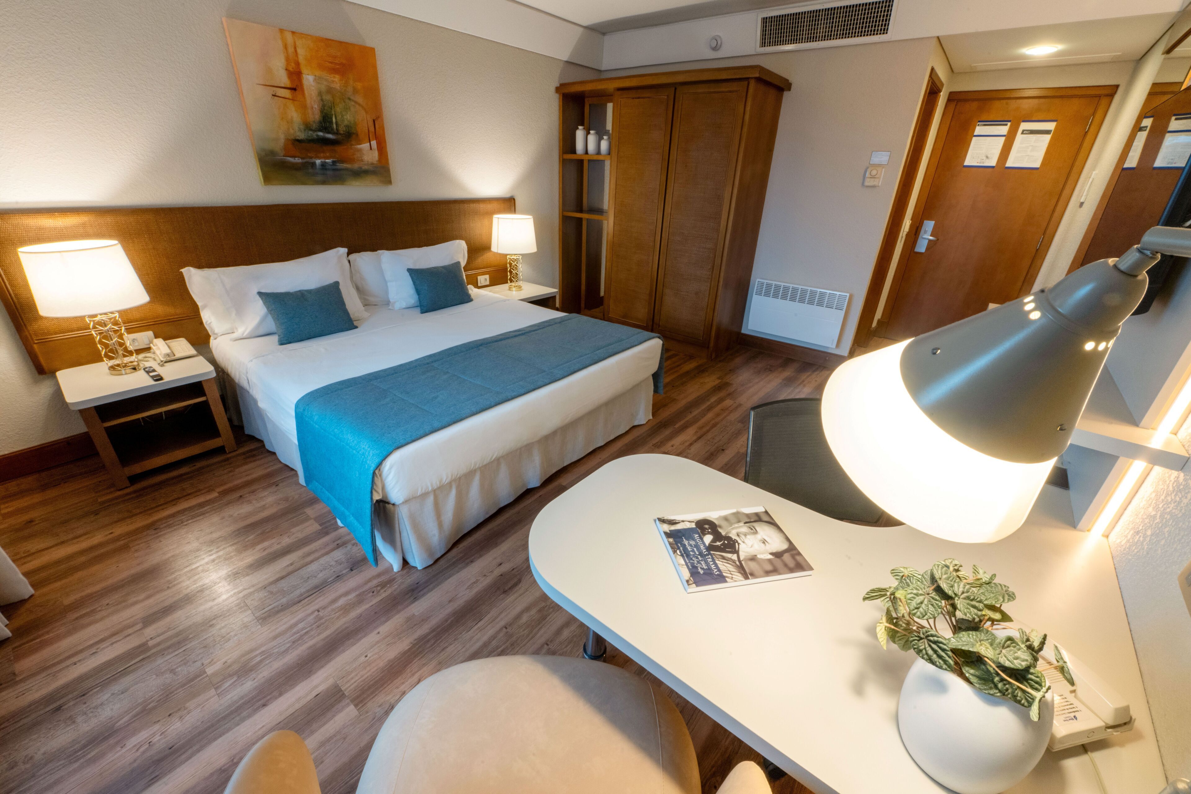 A hotel room with a bed, bedside tables, a desk, a chair, a lamp, a plant, and a wardrobe.