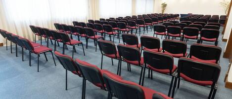 Un salón de conferencias con filas de sillas negras con cojines rojos, piso alfombrado y grandes ventanales con cortinas.