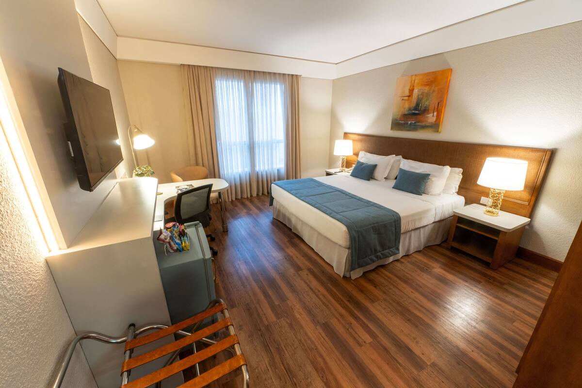A hotel room with a bed, desk, chair, TV, and artwork on the wall.