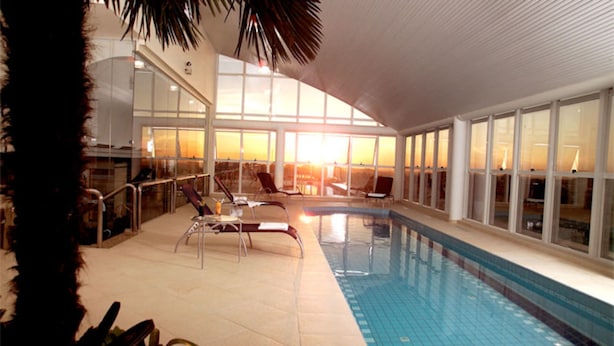 An indoor swimming pool with clear glass walls, lounge chairs, and a view of the sunset through large windows. - Blue Tree Towers Caxias do Sul (Caxias do Sul)