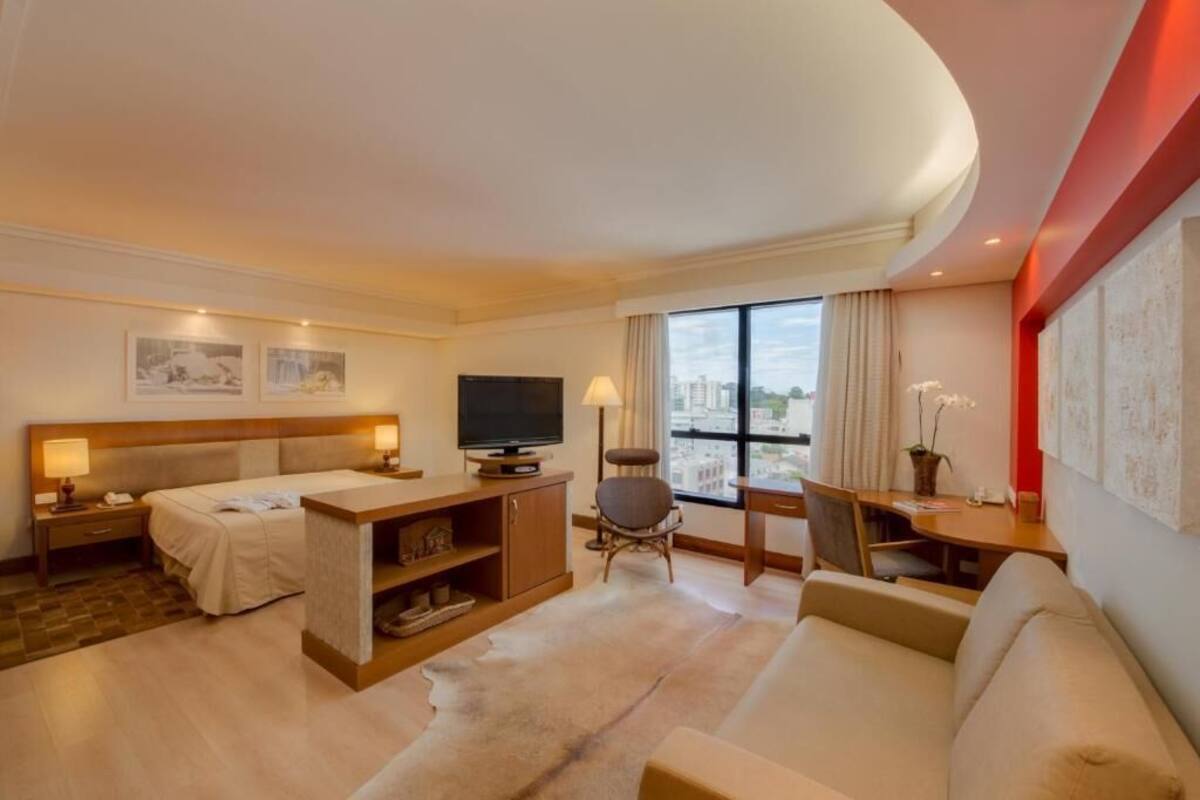 A hotel room with a bed, a desk, a chair, a sofa, a TV, and a large window with a city view.