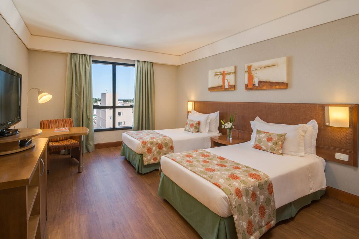 A hotel room with two beds, a desk, a TV, and a window with curtains.