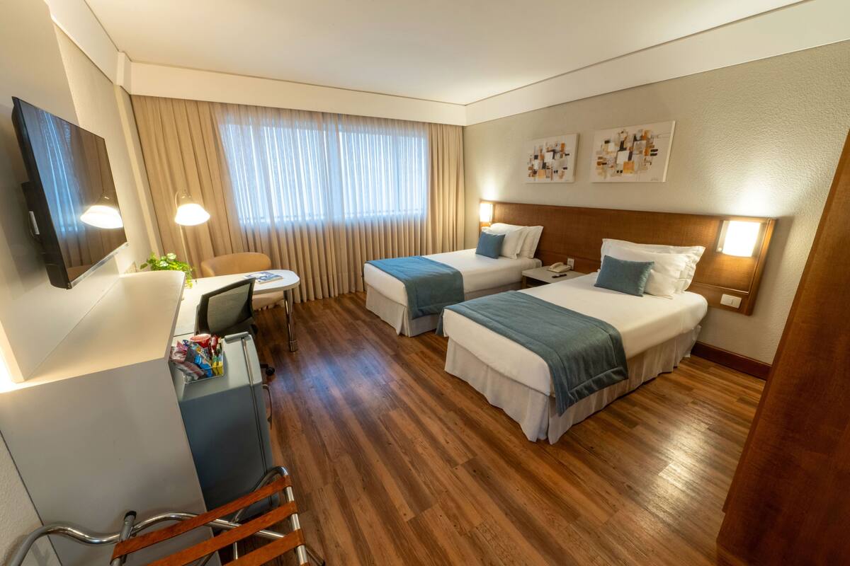 A hotel room with two beds, a desk, a chair, a TV, and a window with curtains.