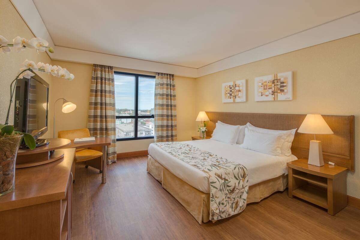 A hotel room with a bed, bedside tables, a desk, a chair, a window with curtains, and artwork on the walls.