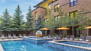 Un área de piscina con sillones de descanso y sombrillas, rodeada de árboles y un edificio con fachada de ladrillo y vidrio.