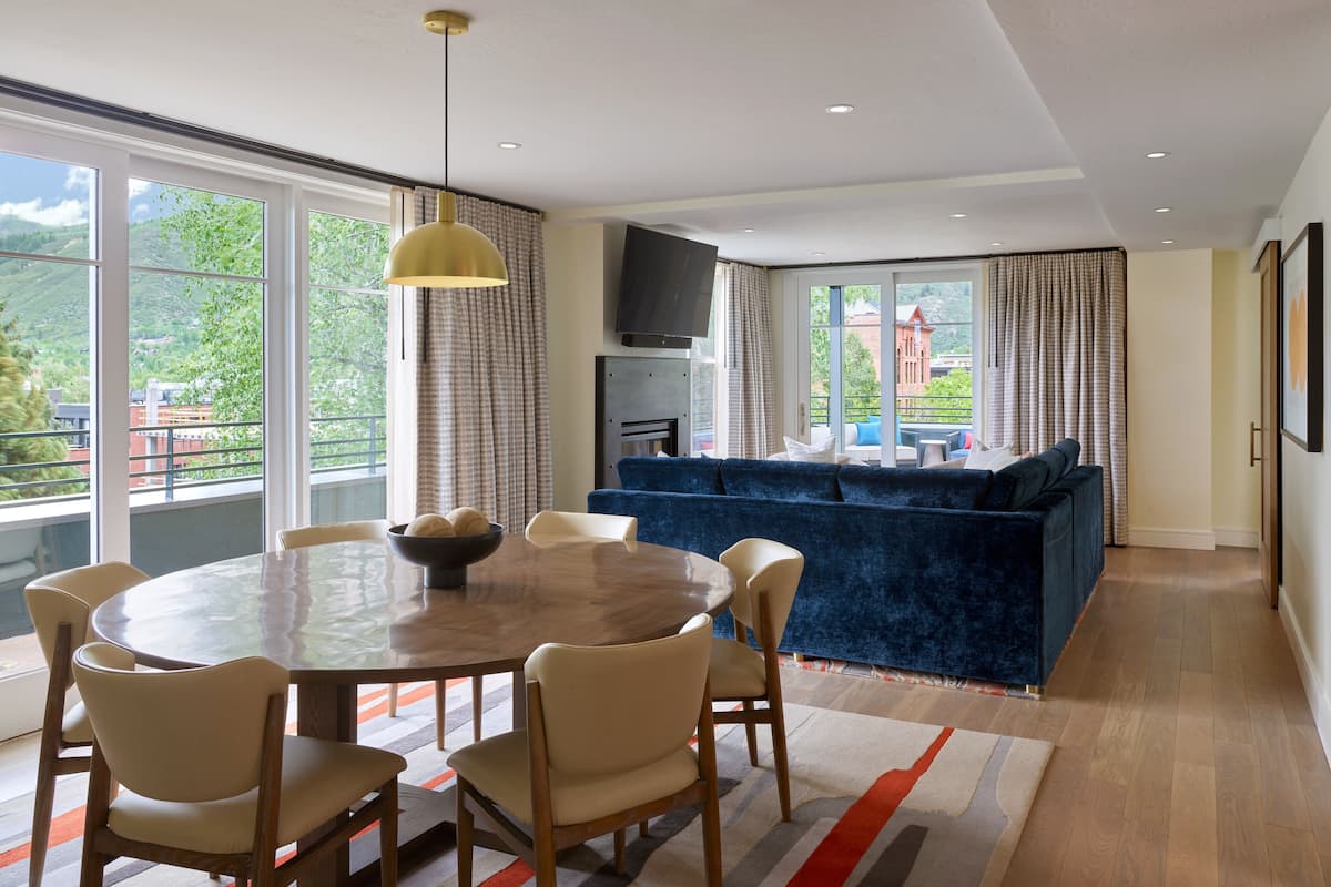 Sala de estar moderna com mesa de jantar redonda, sofá azul e janelas amplas com vista para áreas verdes e edifícios.
