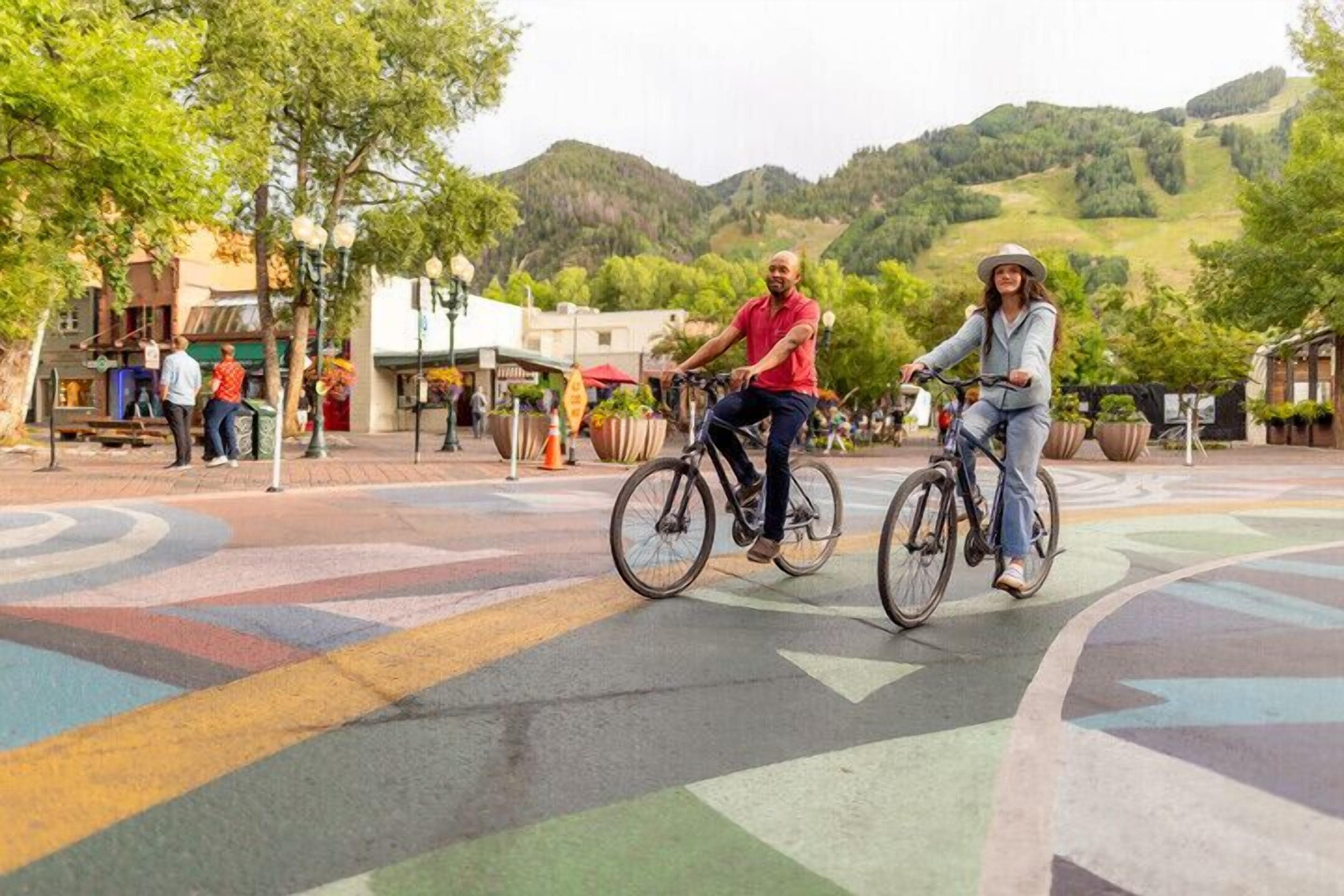 Two people riding bicycles on a colorful, patterned street with shops and trees in the background.