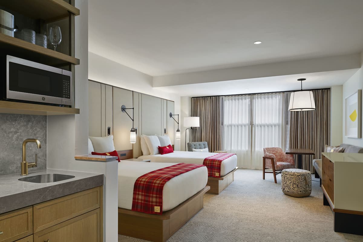 Quarto de hotel com duas camas, uma escrivaninha, uma cadeira, um sofá, um abajur, micro-ondas e pia.