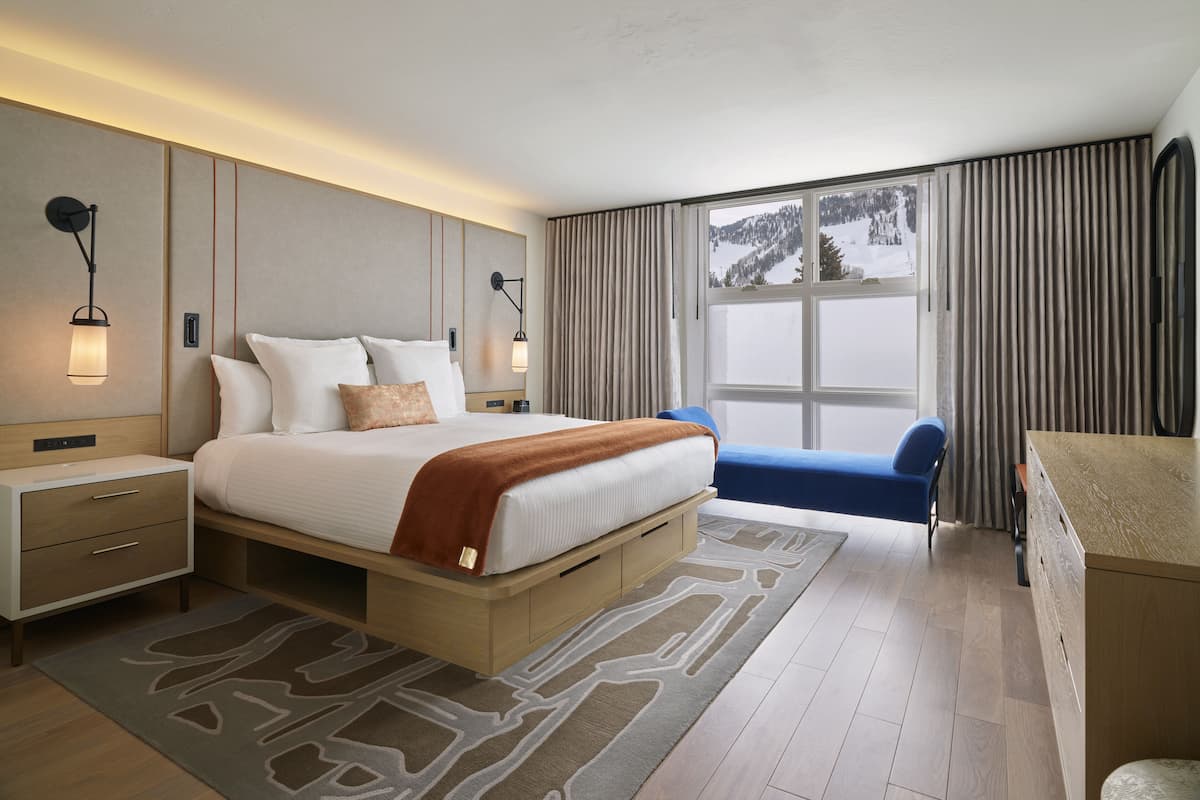 Quarto de hotel moderno com uma cama grande, mesas de cabeceira, escrivaninha e vista para uma paisagem nevada.