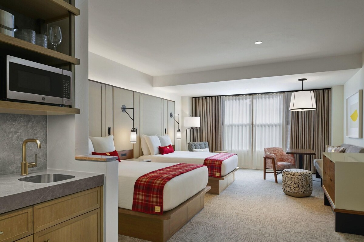 A hotel room with two beds, a desk, a chair, a sofa, a small ottoman, a microwave, a sink, and a minibar.