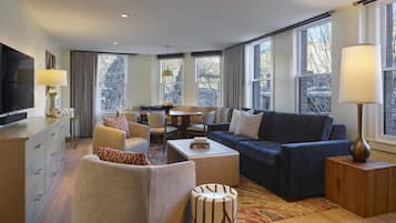 A modern living room with a sofa, armchairs, a coffee table, and a TV. There are large windows with curtains, a wooden floor, and a patterned rug.
