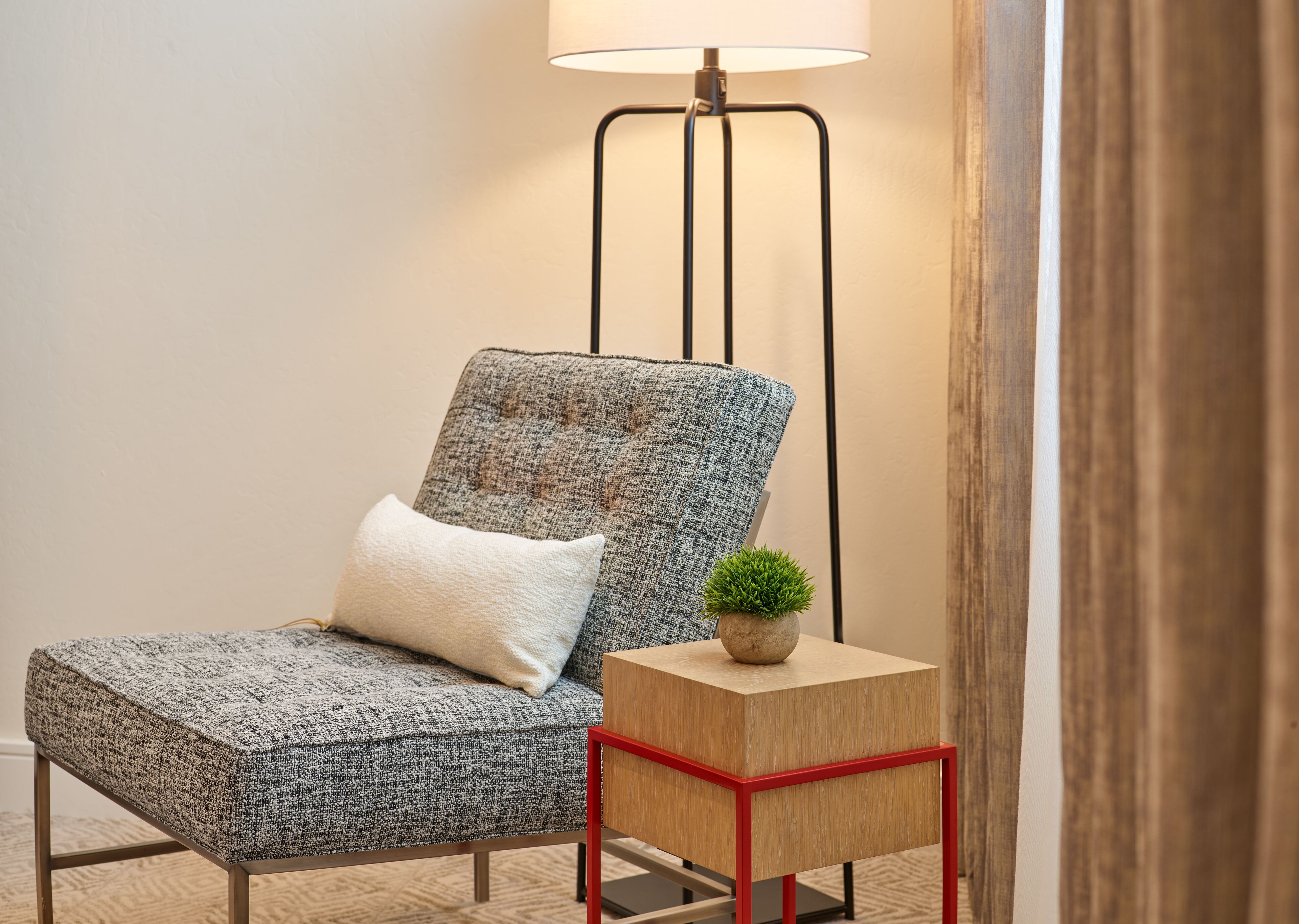 A modern armchair with a textured fabric, a white pillow, and a small potted plant on a wooden side table with red metal legs.
