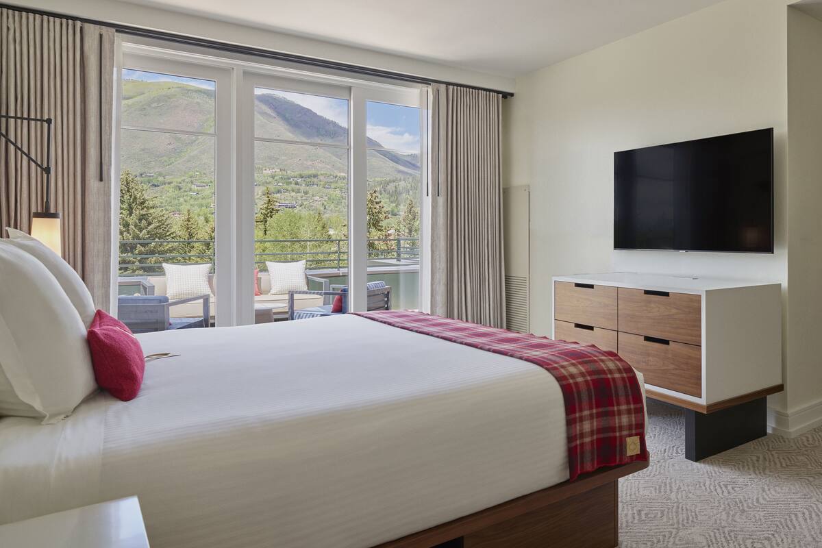 Quarto de hotel com uma cama grande, televisão na parede e varanda com vista para as montanhas.