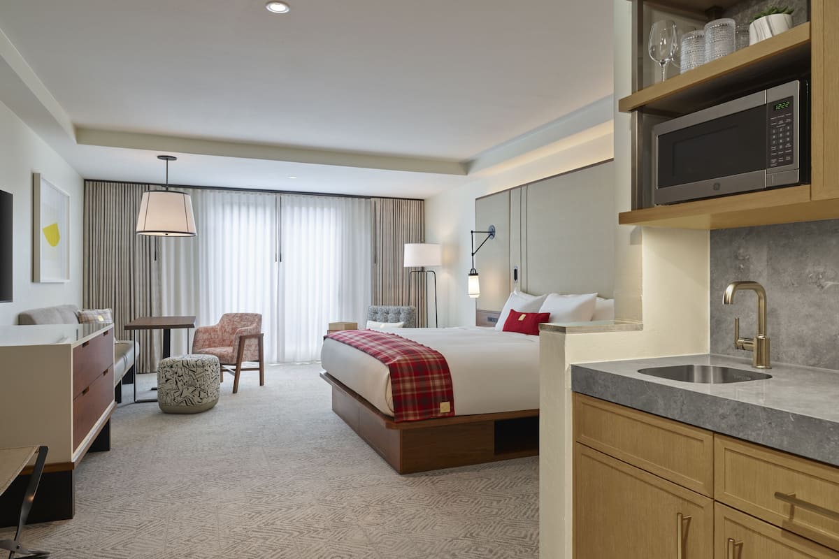Quarto de hotel com cama, escrivaninha, cadeira, micro-ondas, pia e uma janela grande com cortinas.