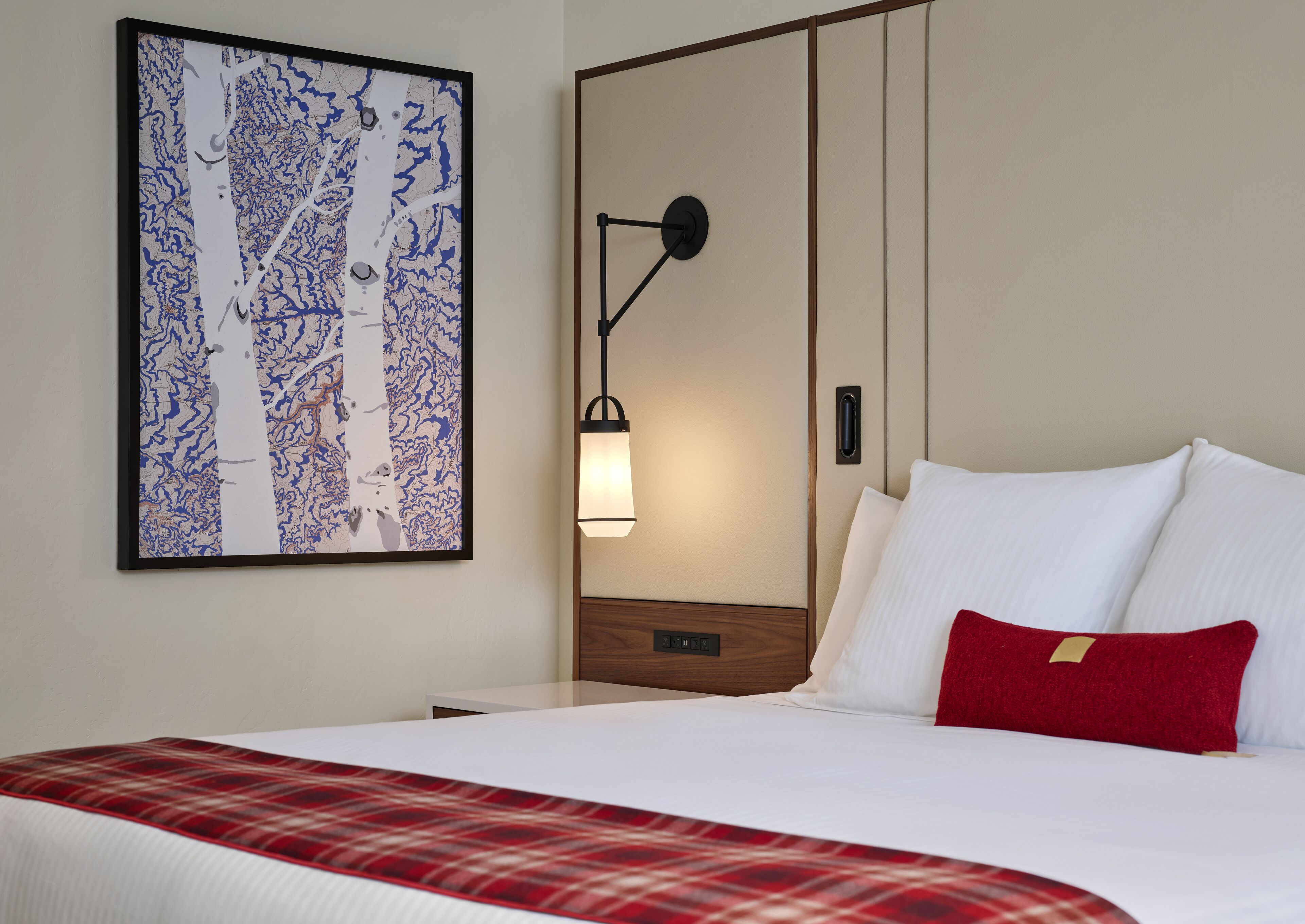 A hotel room with a bed, a red plaid throw blanket, a white pillow, a red pillow, a wall-mounted lamp, and a framed abstract painting.