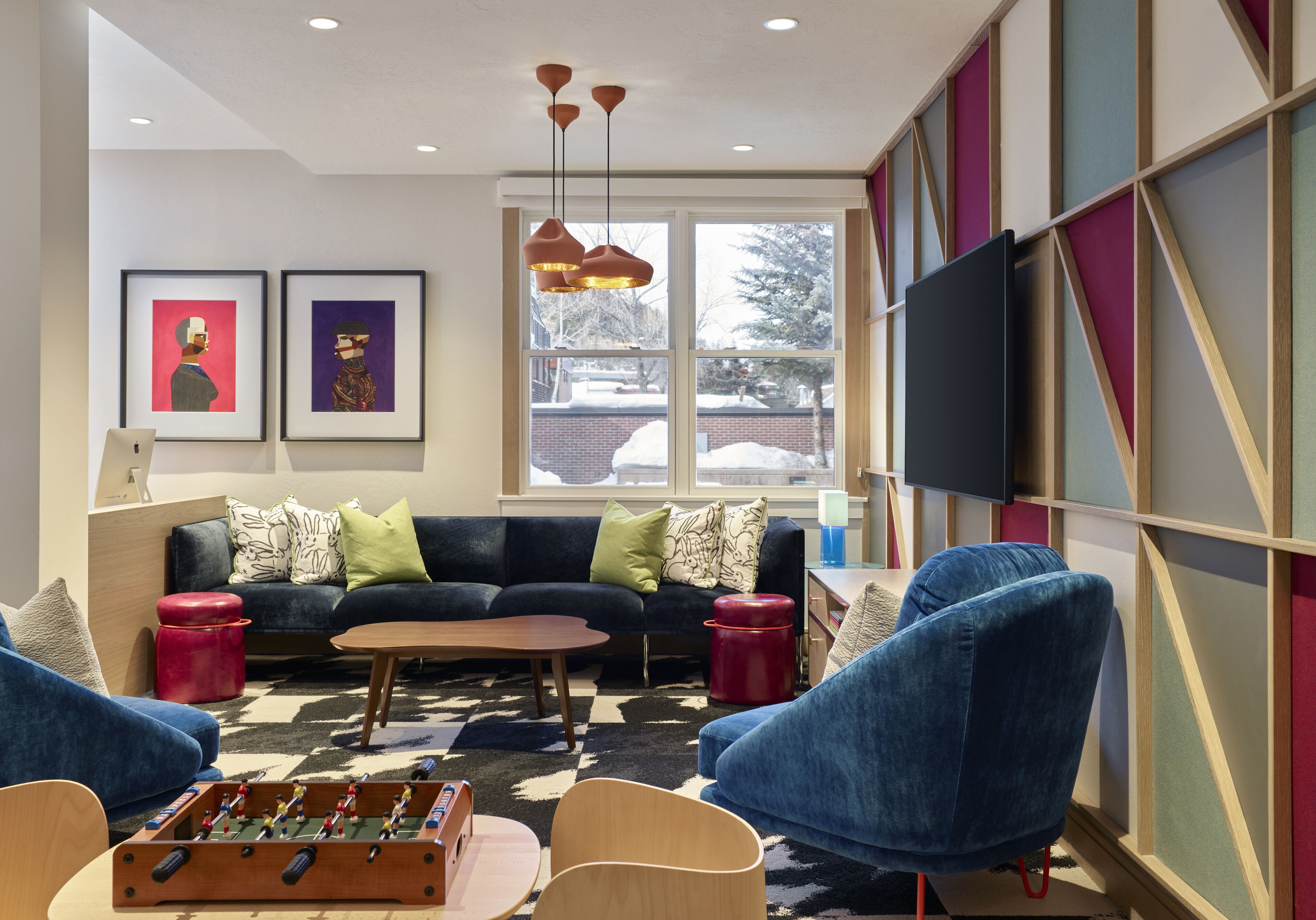 A modern living room with a sofa, chairs, a coffee table, and a foosball table. There is a TV mounted on the wall and a window with a view of the outside.