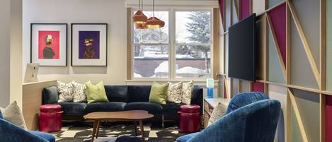 A modern living room with a sofa, chairs, a coffee table, and a foosball table. There is a TV mounted on the wall and a window with a view of the outside.