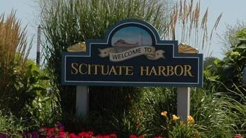 Señal de bienvenida a Scituate Harbor con un paisaje costero y flores.