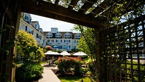 A garden patio area with outdoor seating and umbrellas, surrounded by lush greenery and a pathway leading to a building with multiple windows. - The Inn At Scituate Harbor (Scituate)