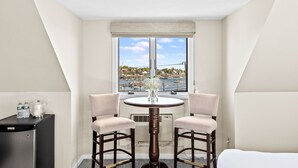 A hotel room with a dining table, two chairs, a small refrigerator, and a window with a view of a marina and buildings. - The Inn At Scituate Harbor (Scituate)