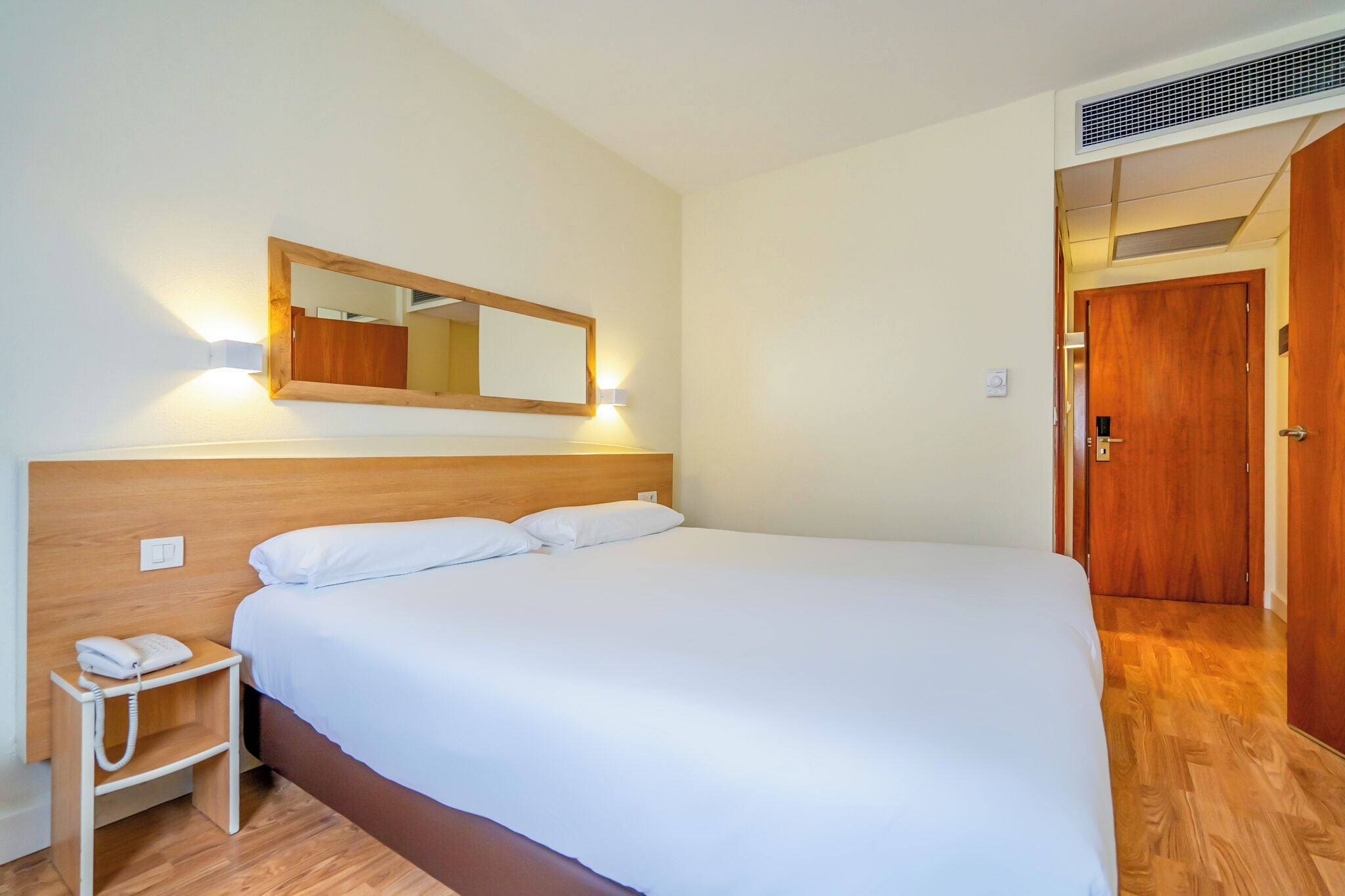 Una habitación de hotel con una cama grande, una mesita de noche con teléfono, un espejo montado en la pared y una puerta de madera.