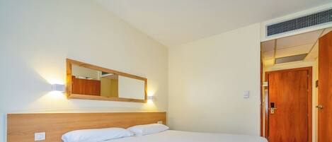 A hotel room with a large bed, a bedside table with a phone, a wall-mounted mirror, and a wooden door.