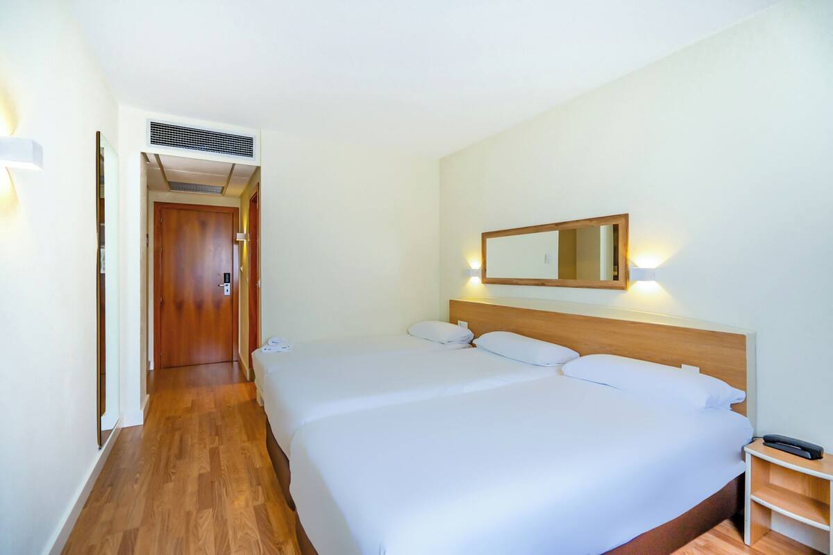 A hotel room with a large bed, a wooden headboard, a bedside table with a lamp, and a mirror on the wall.