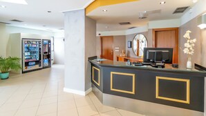 A hotel reception area with a black reception desk, a vending machine, a potted plant, and a decorative wall piece. - Hotel YIT Ciudad de Elche (Elche)