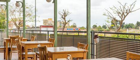 An outdoor seating area with wooden tables and chairs, a green metal fence, and a view of buildings and trees in the background.