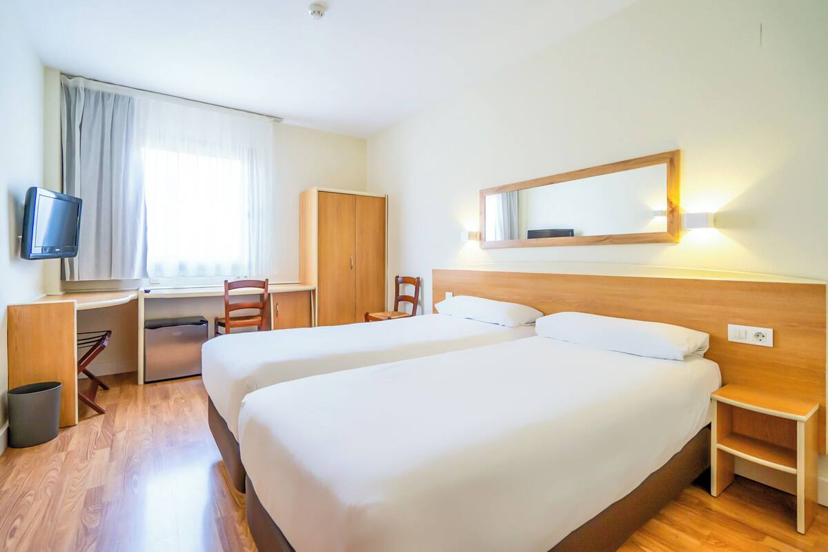 A hotel room with two beds, a desk, a chair, a TV, and a wardrobe.